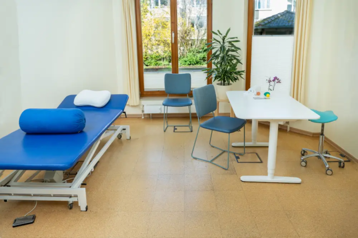 Therapiezimmer mit blauer Liege, einem Tisch und Stühlen.